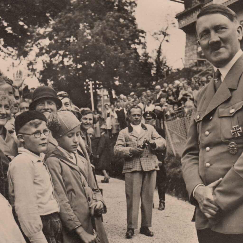 Schwarz-weiß Foto, das rechts Adolf Hitler, in der Mitte Heinrich Hoffmann mit Kamera und links Kinder zeigt